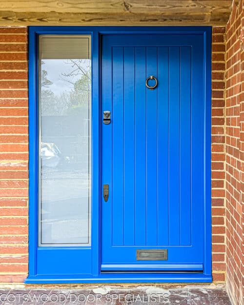 Blue contemporary front door with sidelight frame . Door is made in a tradional way with vertical boards. Frame has a full height piece of glass that is sandblasted with a clear inset line border. Door has dark bronze banham locks, letterplate and knocker. Door and frame painted blue