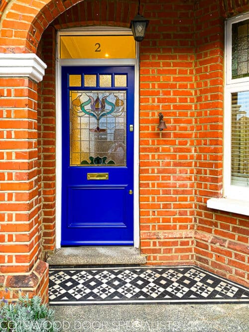 Floral Edwardian front door. Painted blue Edwardian front door with elegant floral design stained glass. Polished brass door hardware. Door is fitted into a red brick house. Stained glass has different textures and a reeded border.