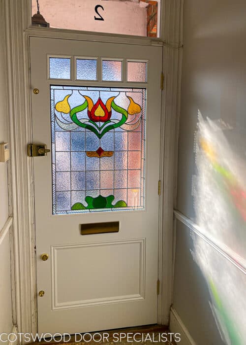 Floral Edwardian front door. Painted blue Edwardian front door with elegant floral design stained glass. Polished brass door hardware. Door is fitted into a red brick house. Stained glass has different textures and a reeded border. Internal hallway photo showing off stained glass