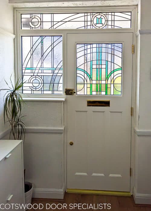 Art Deco front entrance. 1930s front door fitted in london with geometric Jean Dunand inspired stained glass. Glass uses lots of different textures with some flashes of coloured teal glass. The glass has a number above the door in coloured glass. Door is painted light blue with a white door frame. Door is fitted in a london brick home and has polished brass door furniture. Interior hallway photo highlighting stained glass