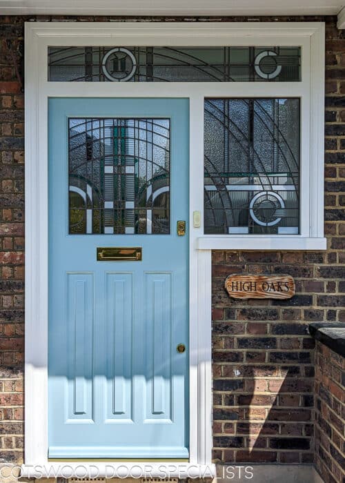 Art Deco front entrance. 1930s front door fitted in london with geometric Jean Dunand inspired stained glass. Glass uses lots of different textures with some flashes of coloured teal glass. The glass has a number above the door in coloured glass. Door is painted light blue with a white door frame. Door is fitted in a london brick home and has polished brass door furniture.