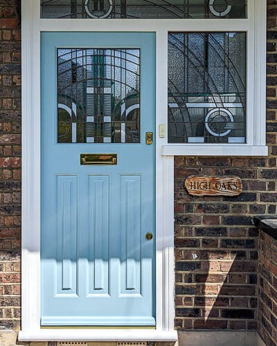 Art Deco front entrance. 1930s front door fitted in london with geometric Jean Dunand inspired stained glass. Glass uses lots of different textures with some flashes of coloured teal glass. The glass has a number above the door in coloured glass. Door is painted light blue with a white door frame. Door is fitted in a london brick home and has polished brass door furniture.