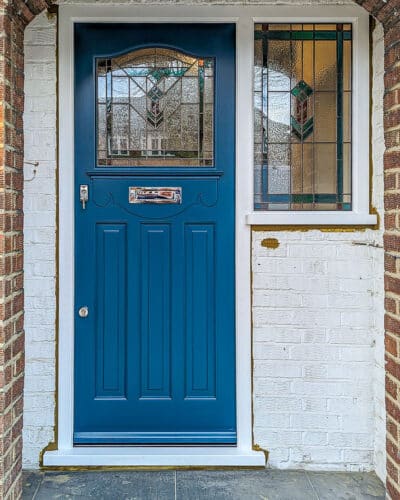 Leaded light 1930s Door. 1930s front door with wing window door frame. Door and frame made in Accoya wood and painted in Teknos paint. Door is painted blue and the door frame is painted white. Door furniture is polished chrome and the door has a letterplate with a knocker on it. The glass is a bespoke design and is a leadlight with vibrant colours- teals and reads. Different textures add depth to the glass design and the glass is made as a sealed double glazed unit. The stained glass design is a geometric art deco design. The door is a 1930s classic design with 3 panels and a shaped piece of glass