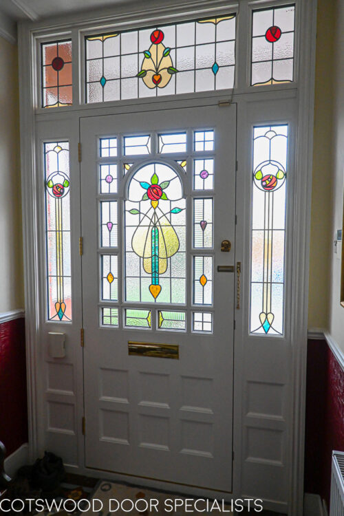 ornamental Edwardian front door with stained glass. A decorative 17 light Edwardian front door with decorative features such as a shelf and dentil block and an apron of which the letterplate is fixed too. The frame is a large frame with sidelights and glazing above the door. The door frame is very ornamental with pilasters (detailed moulding forming columns each side of the door) supporting a decorative transom with detailed mouldings and a dentil block. The stained glass is a art nouveau design featuring roses. door and frame are a replica of the orignal Edwardian joinery. Handmade in Accoya wood and fitted with multipoint locking. Internal picture of stained glass