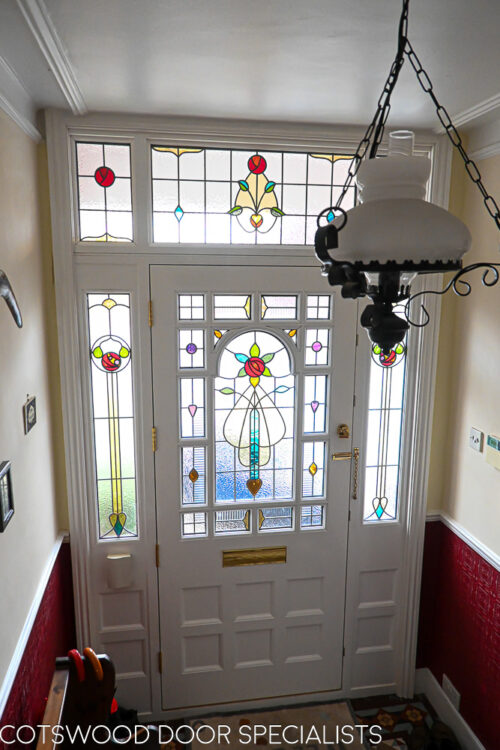 ornamental Edwardian front door with stained glass. A decorative 17 light Edwardian front door with decorative features such as a shelf and dentil block and an apron of which the letterplate is fixed too. The frame is a large frame with sidelights and glazing above the door. The door frame is very ornamental with pilasters (detailed moulding forming columns each side of the door) supporting a decorative transom with detailed mouldings and a dentil block. The stained glass is a art nouveau design featuring roses. door and frame are a replica of the orignal Edwardian joinery. Handmade in Accoya wood and fitted with multipoint locking. Internal picture of stained glass in hallway