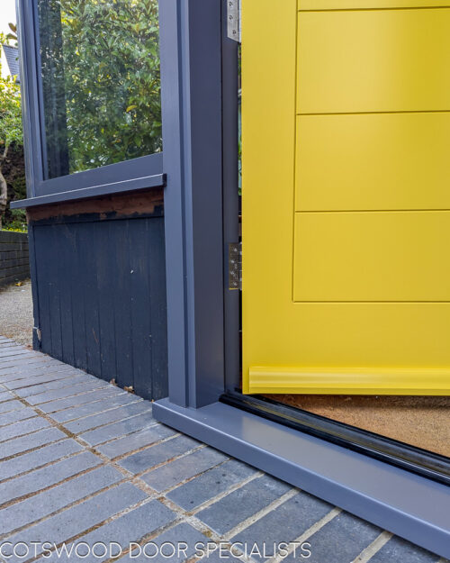 Yellow Contemporary front door. New accoya wooden door fitted into a new wooden frame. Door is contemporary in style with lots of glazing surrounding the door. The frame is painted grey and the door a bright yellow. The door is solid wood with no glass and horizontal wooden panels. Satin chrome door furniture and door locks. Closeup of panel and draught proofing