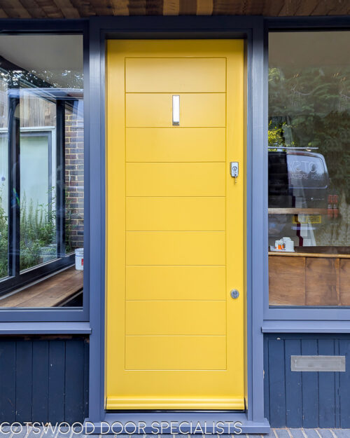 Yellow Contemporary front door. New accoya wooden door fitted into a new wooden frame. Door is contemporary in style with lots of glazing surrounding the door. The frame is painted grey and the door a bright yellow. The door is solid wood with no glass and horizontal wooden panels. Satin chrome door furniture and door locks